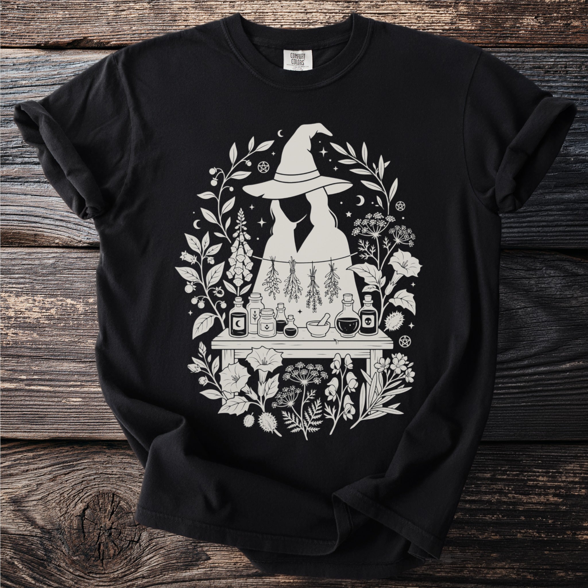 Black t-shirt with a light-colored witchy potion-making graphic and floral accents.