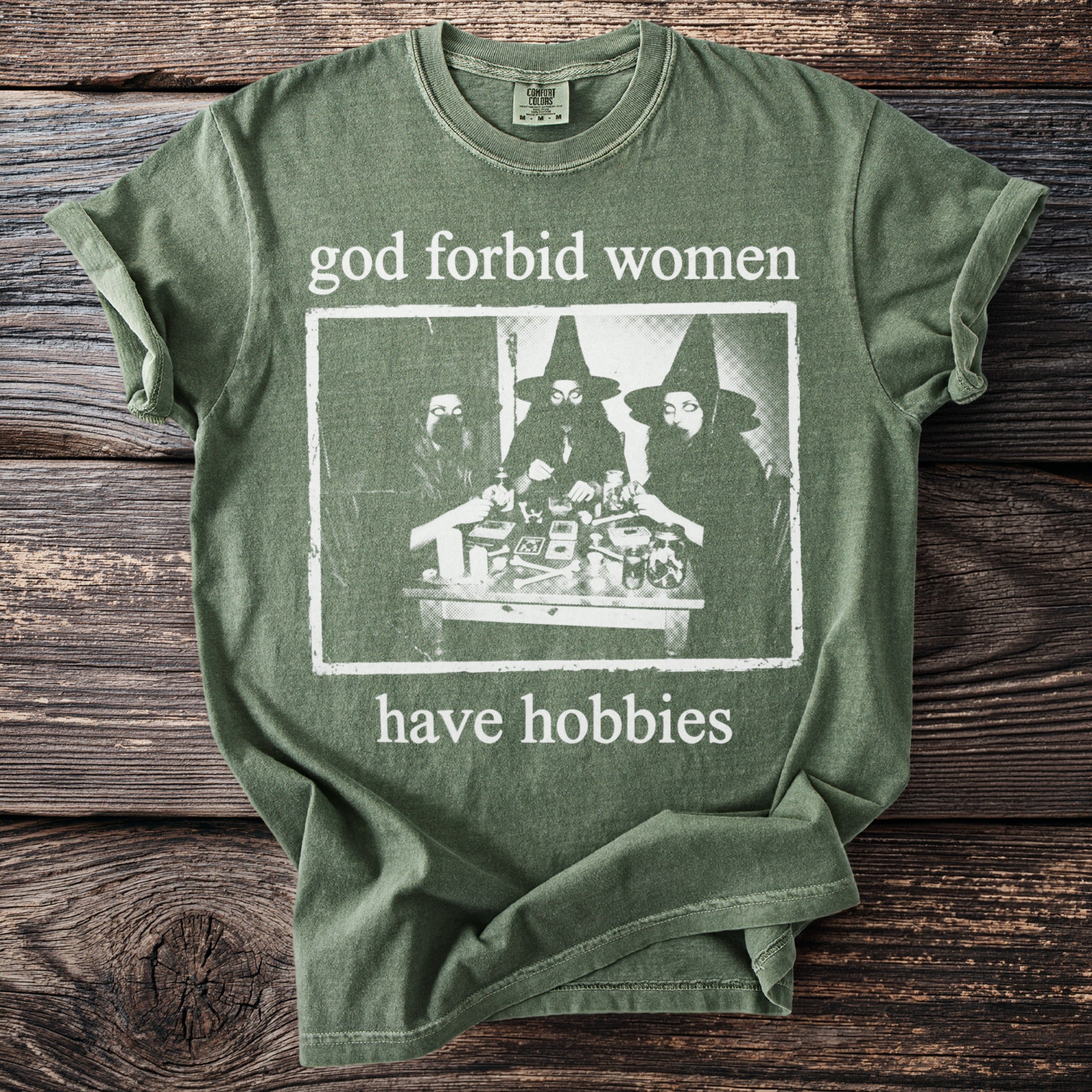Green t-shirt with text "god forbid women have hobbies" and image of three people in witch hats.