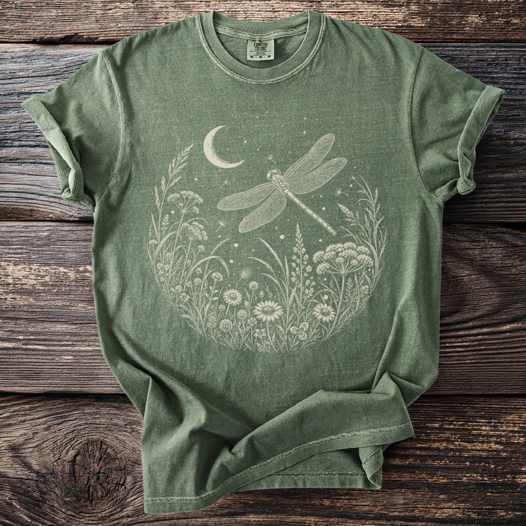 Green t-shirt with dragonfly, moon, and wildflowers graphic on rustic wood.