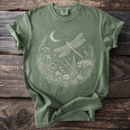 Green t-shirt with dragonfly, moon, and wildflowers graphic on rustic wood.