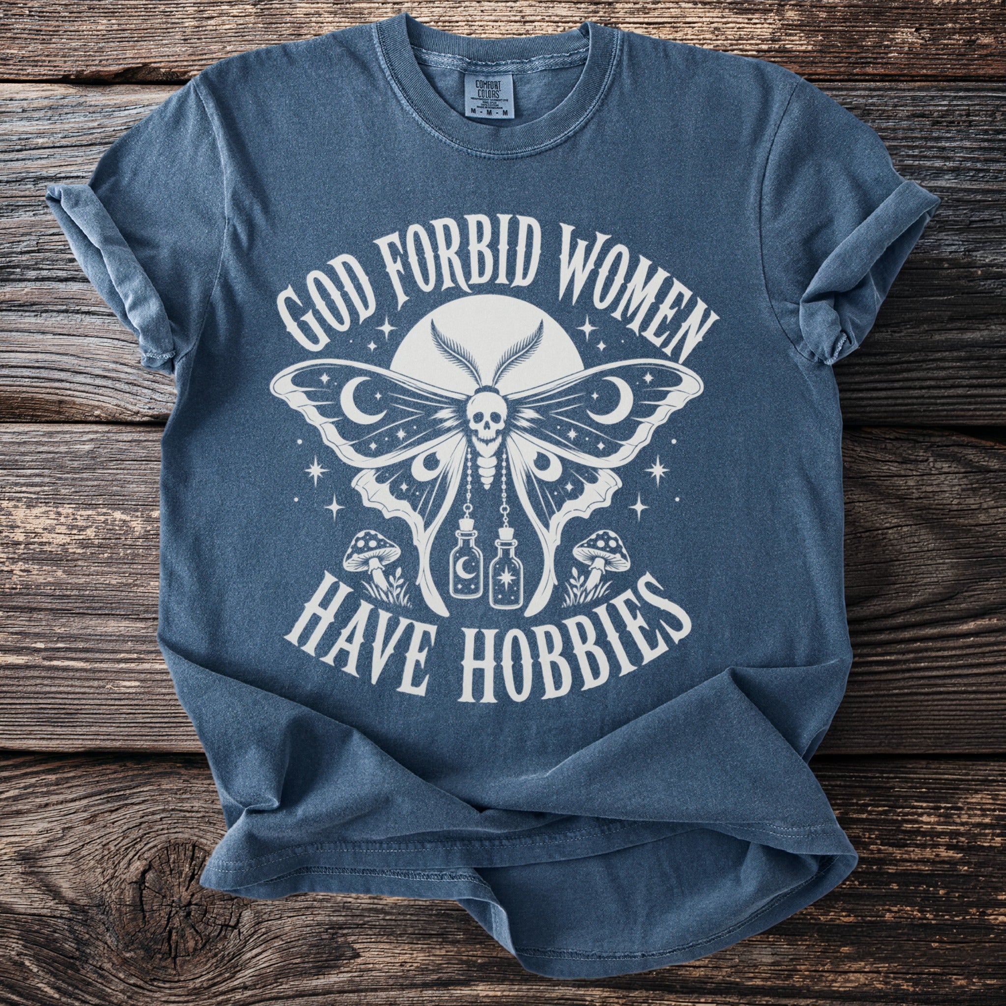 Blue t-shirt with white text: "God Forbid Women Have Hobbies" and moth graphic.