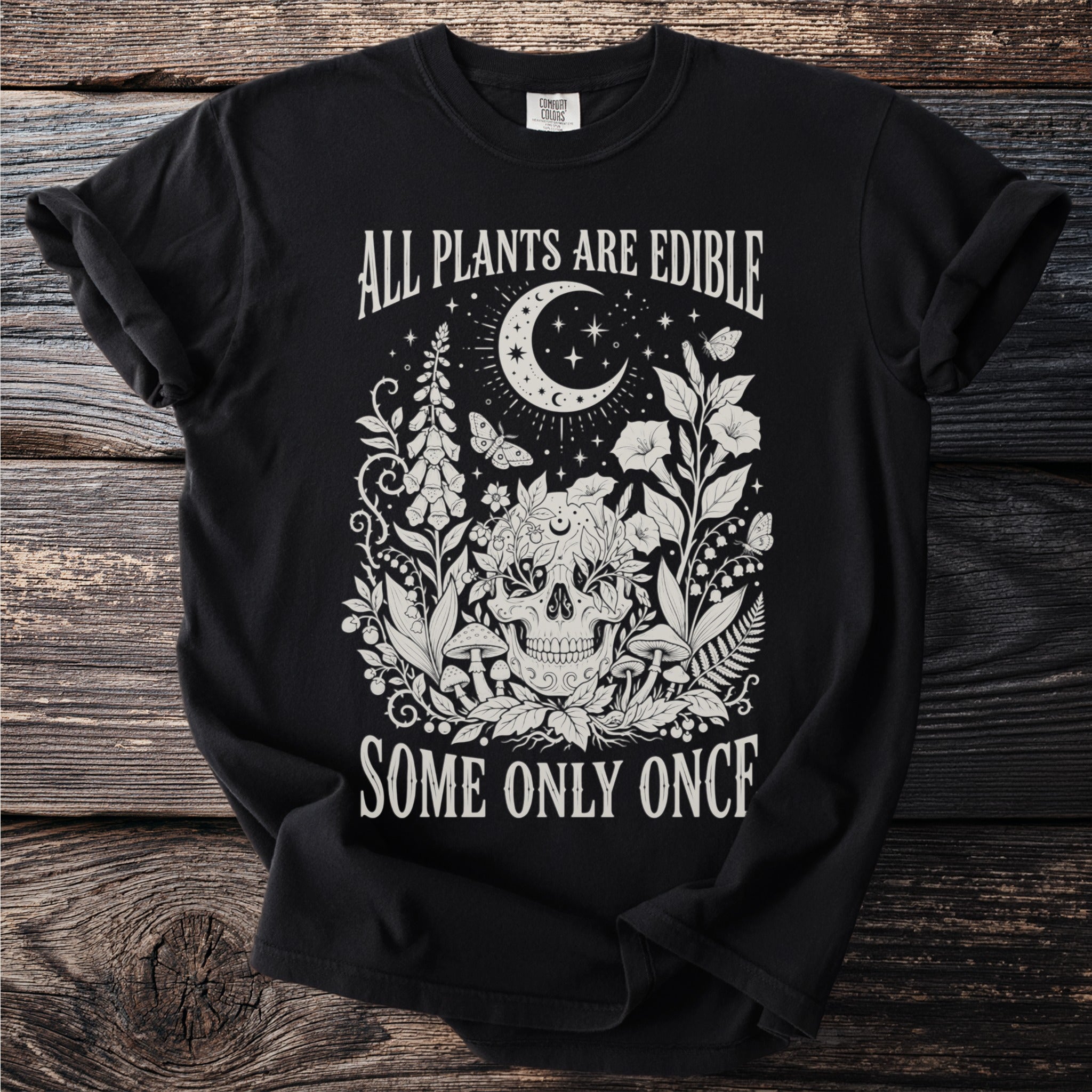 Black t-shirt with a design of a skull surrounded by plants, moon, stars, and butterflies. Text reads "All Plants Are Edible Some Only Once".