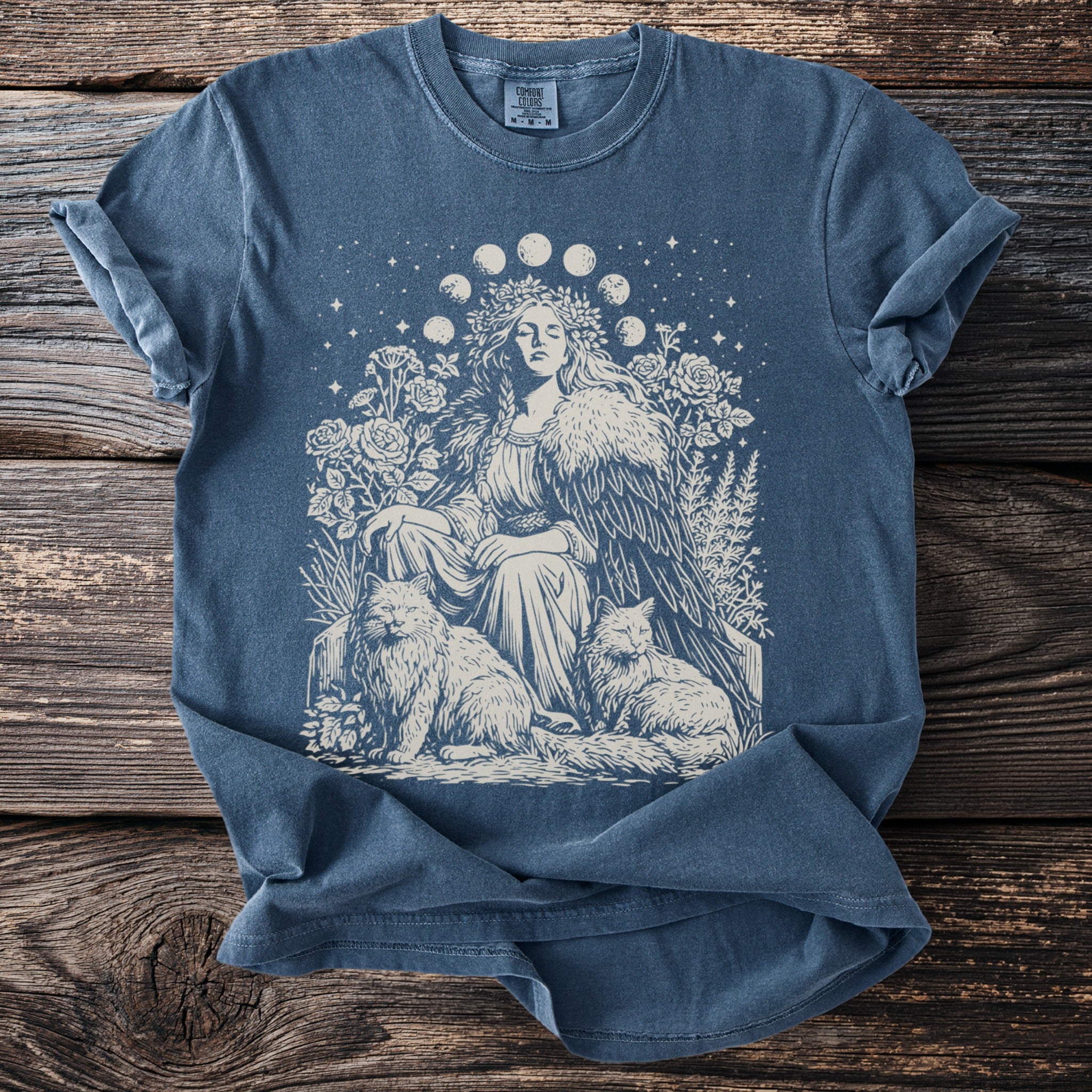 Blue t-shirt with a pale graphic of a woman, cats, and floral elements under a moon phase garland.