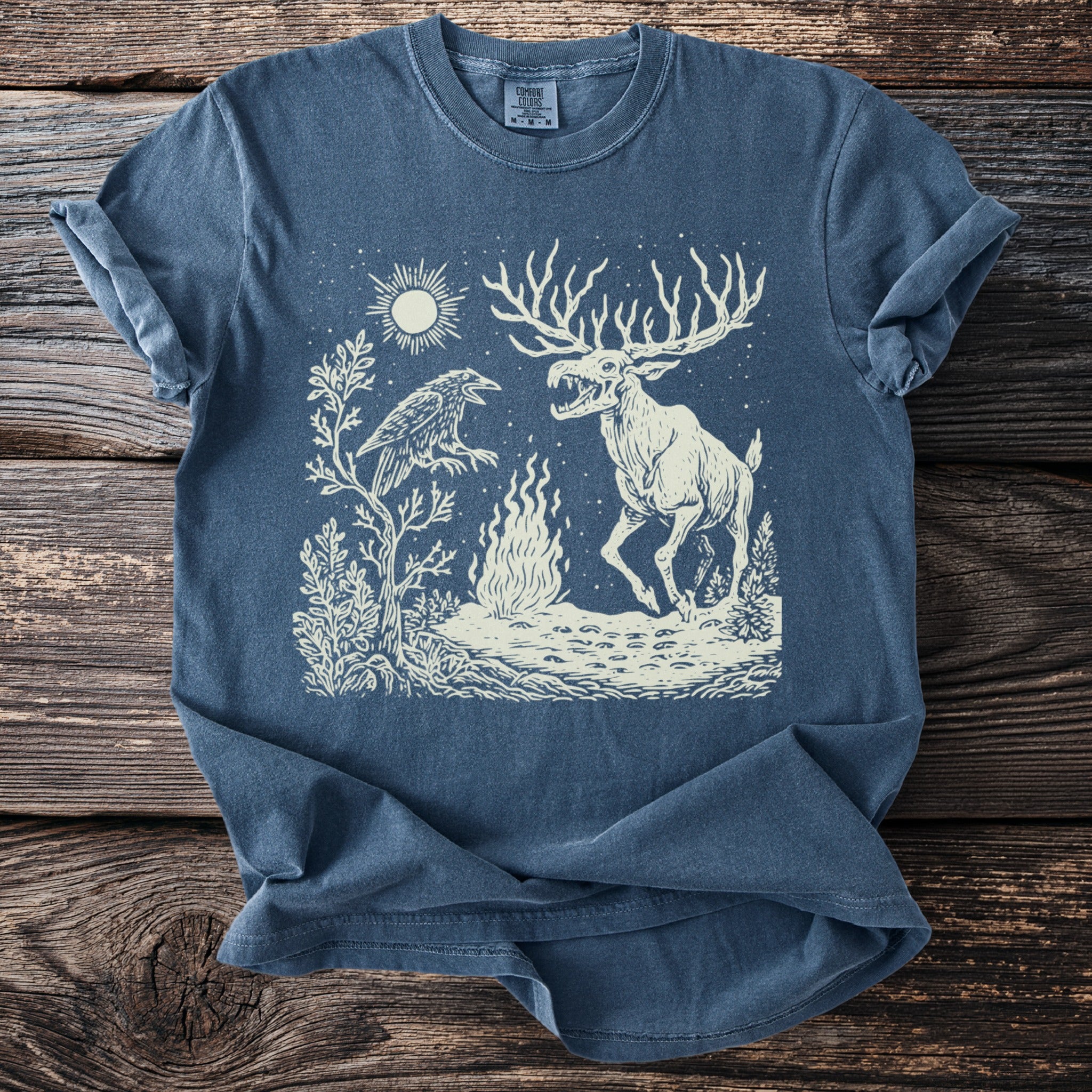 Blue t-shirt with a cream illustration of a raven, a fire, and a monstrous deer under a sun.