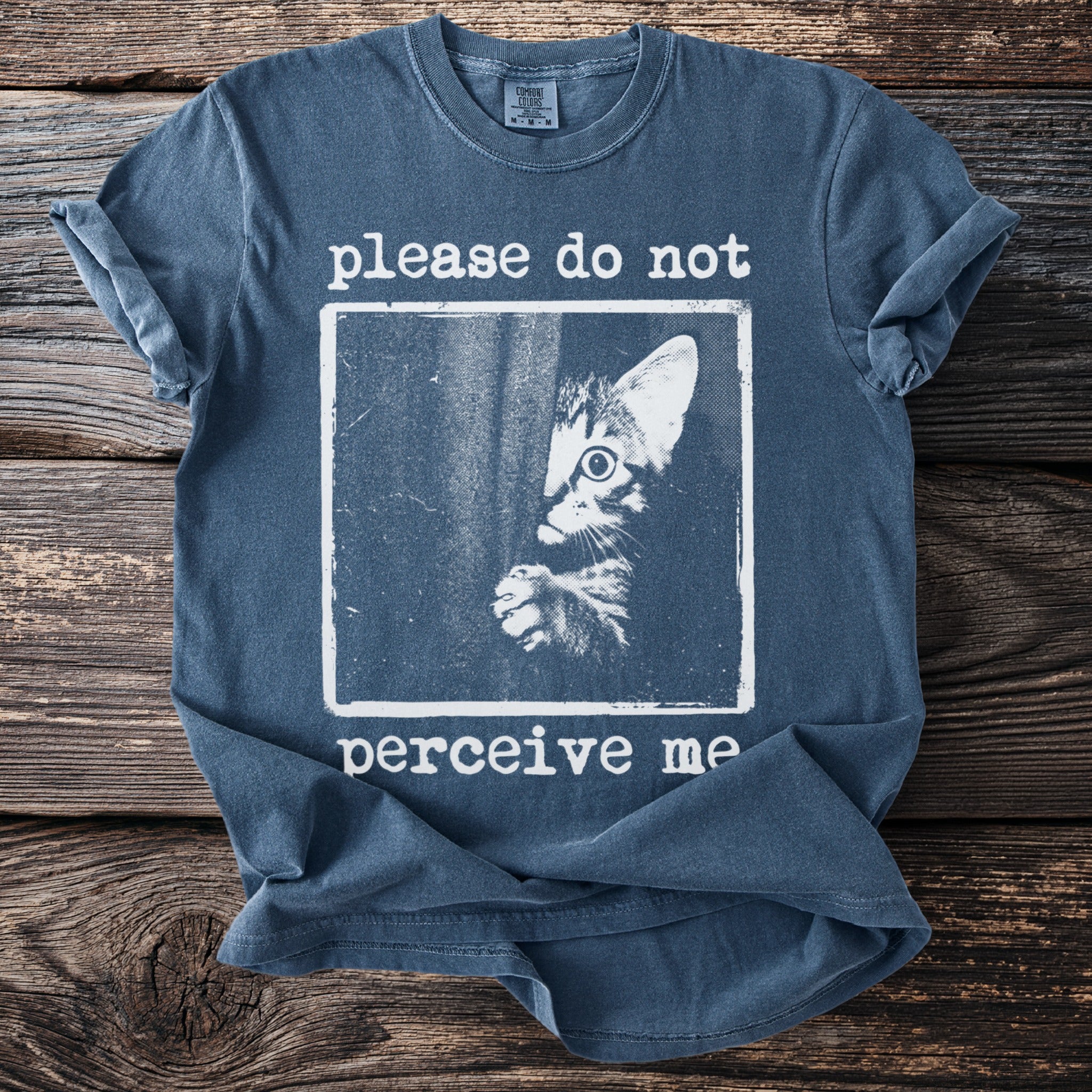 Blue t-shirt with a graphic of a shy cat and text "please do not perceive me".