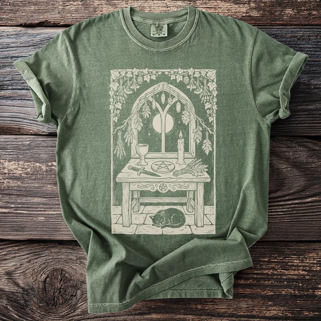 Green t-shirt with a mystical altar design featuring a cat, witchy tools, and a crescent moon in a window.