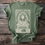 Green t-shirt with a mystical altar design featuring a cat, witchy tools, and a crescent moon in a window.