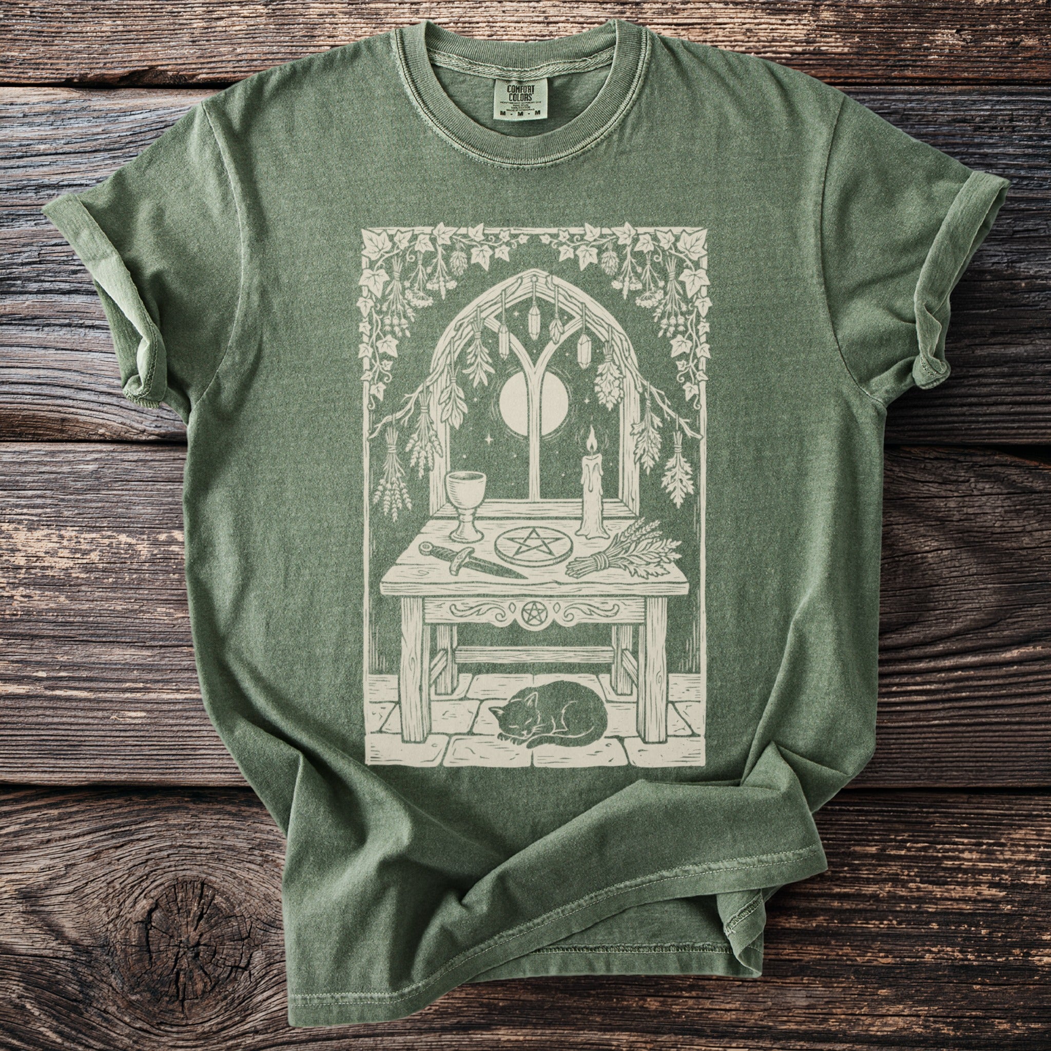 Green t-shirt with a mystical altar design featuring a cat, witchy tools, and a crescent moon in a window.