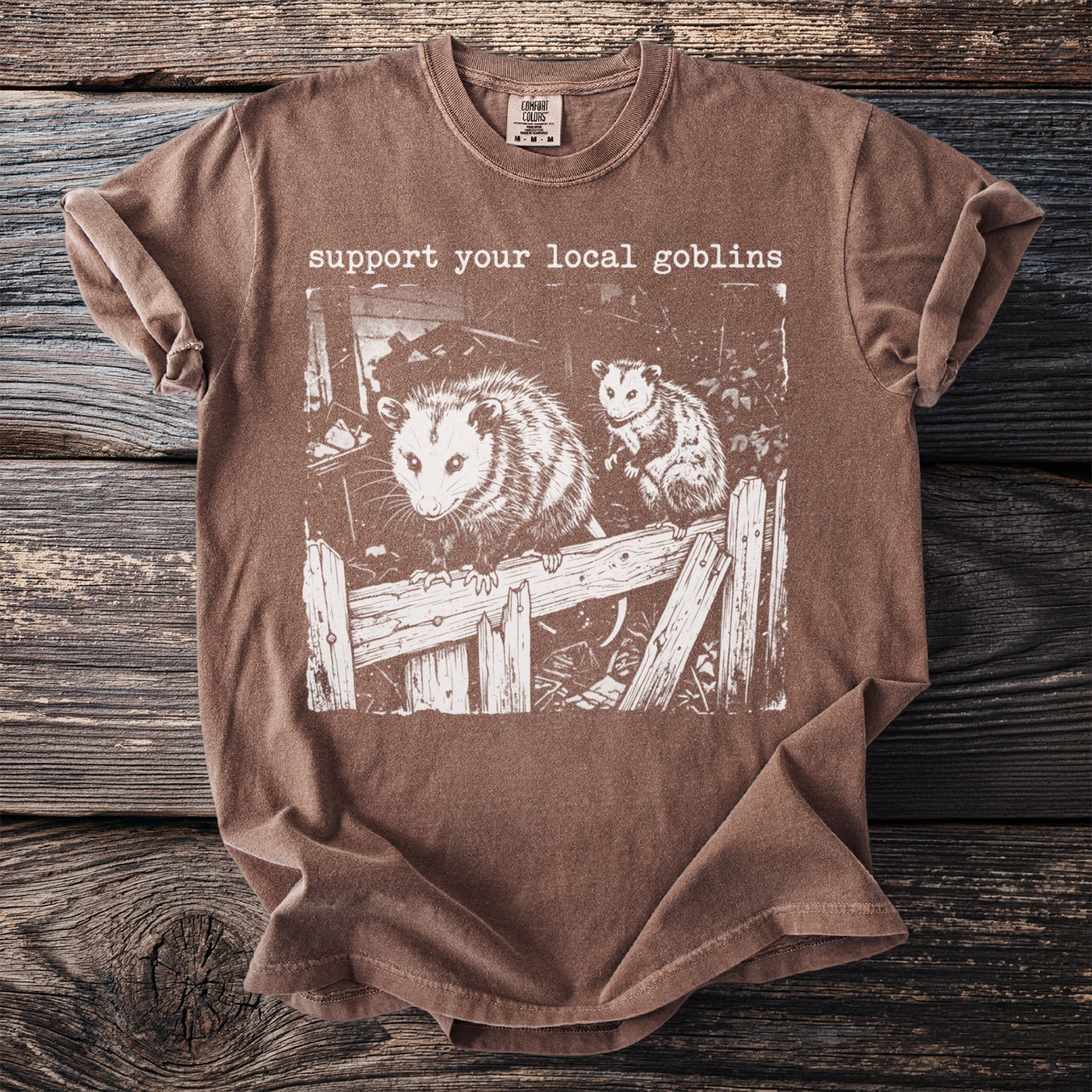 Brown t-shirt with "support your local goblins" and an illustration of two opossums.