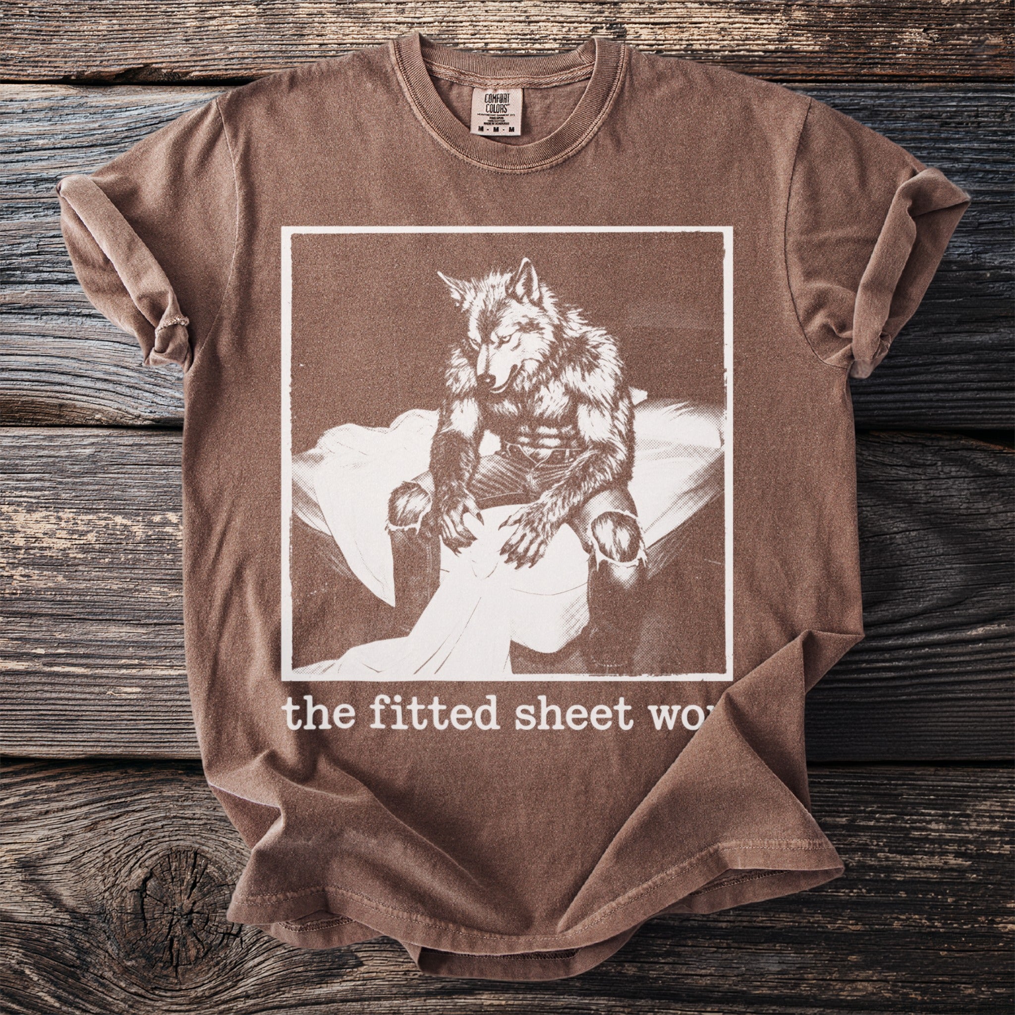 Brown t-shirt with a graphic of a werewolf on a bed and text "the fitted sheet wo".