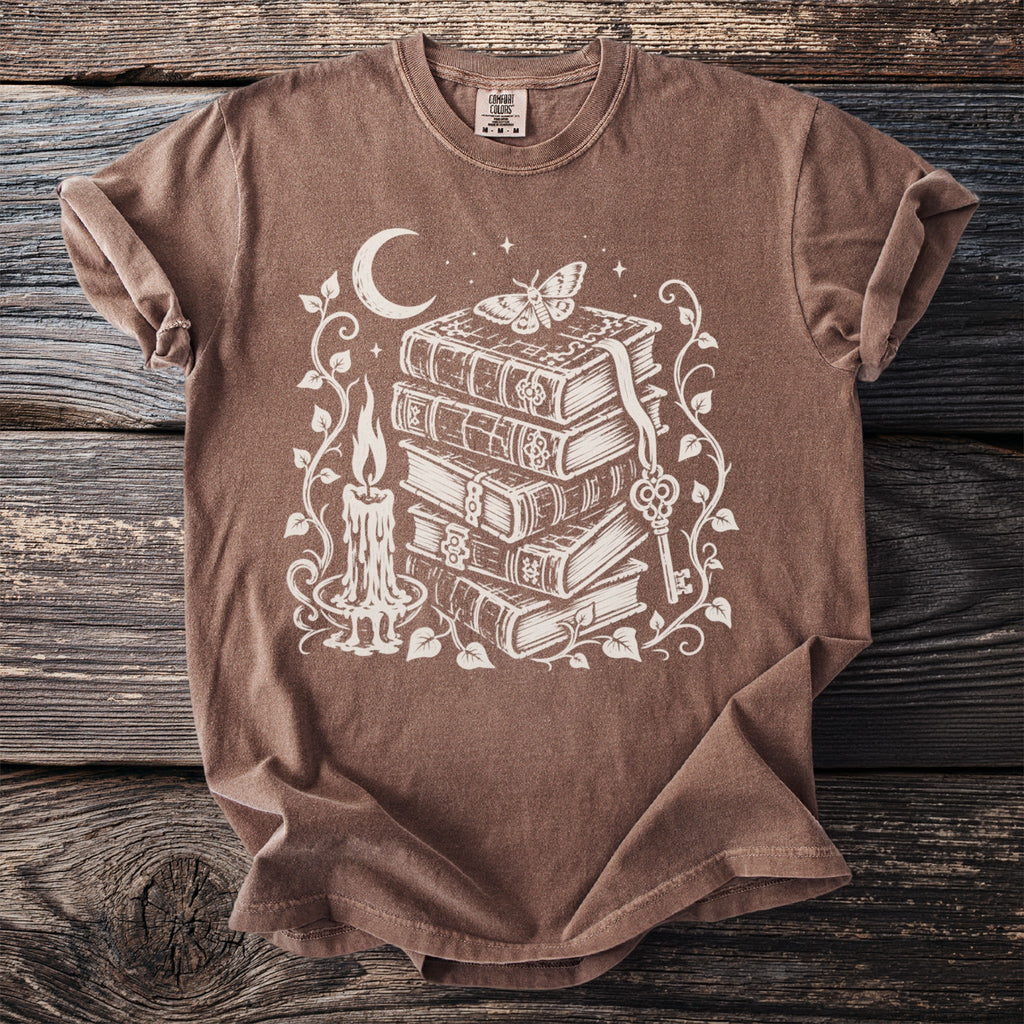 Brown t-shirt with a white graphic of books, candle, moon, key, and moth.