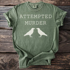 Green t-shirt with "ATTEMPTED MURDER" and two white bird silhouettes.