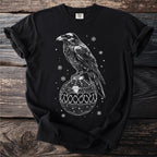 Black t-shirt with white graphic of a raven perched on a decorative ornament with snowflakes.