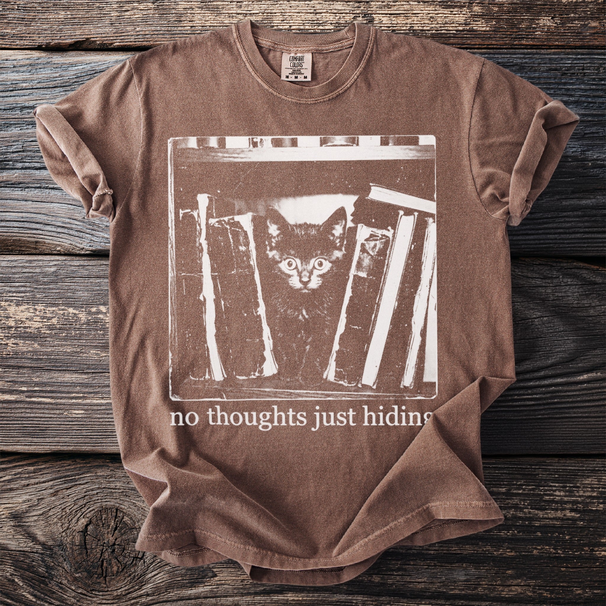 Brown t-shirt with white graphic of kitten hiding among books and text "no thoughts just hiding".