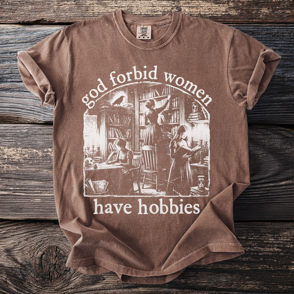 Brown t-shirt with "God forbid women have hobbies" text and library scene graphic.