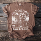 Brown t-shirt with "God forbid women have hobbies" text and library scene graphic.