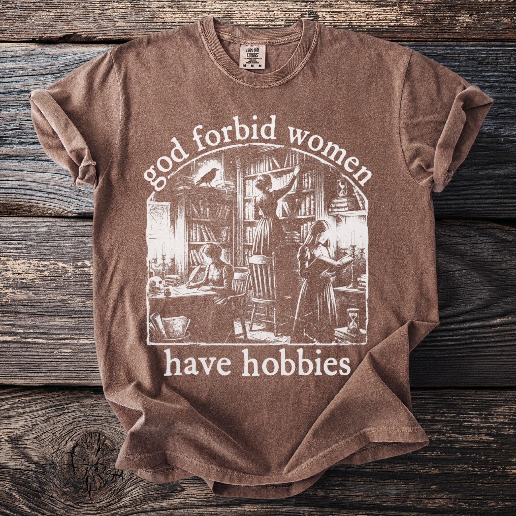 Brown t-shirt with "God forbid women have hobbies" text and library scene graphic.