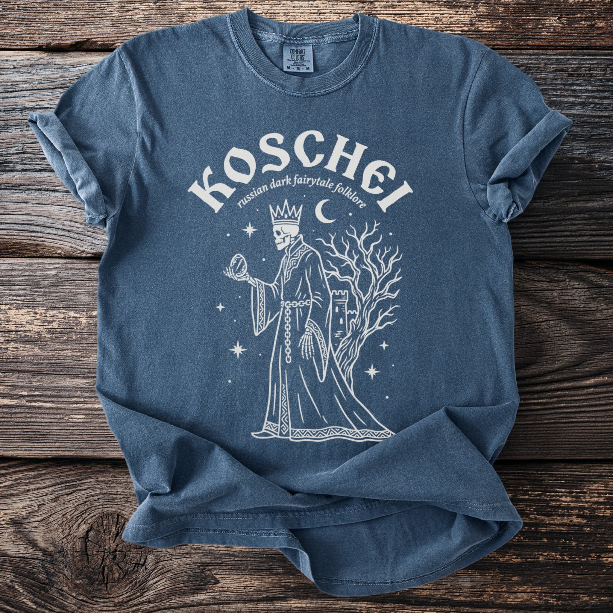 Blue t-shirt with Koschei graphic: skeleton king holding orb, castle, tree, moon, stars.