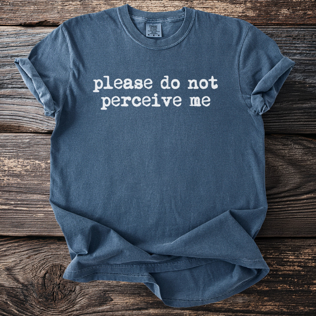 Blue t-shirt with white text "please do not perceive me" on wooden background.