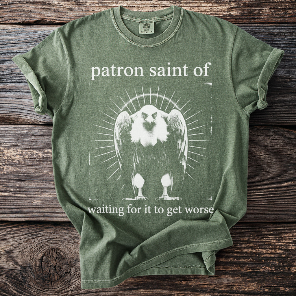 Green t-shirt with white graphic: "patron saint of" above a vulture, with "waiting for it to get worse" below.