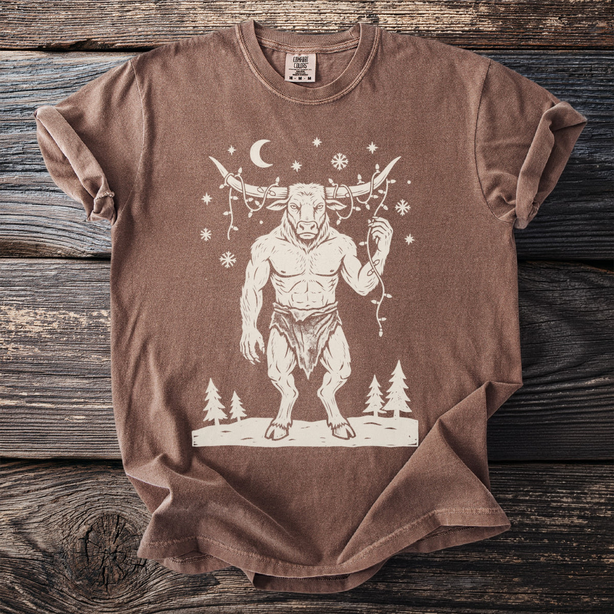Espresso brown t-shirt with a cream folk-cut print: the Minotaur stands in the snow with a string of Christmas lights tangled around his horns.