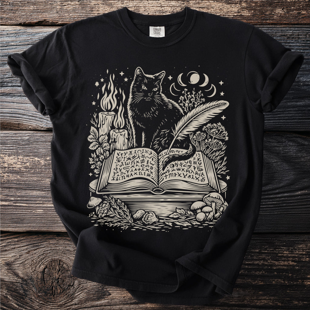 Black t-shirt with a cream graphic of a cat reading an open book with mystical symbols.