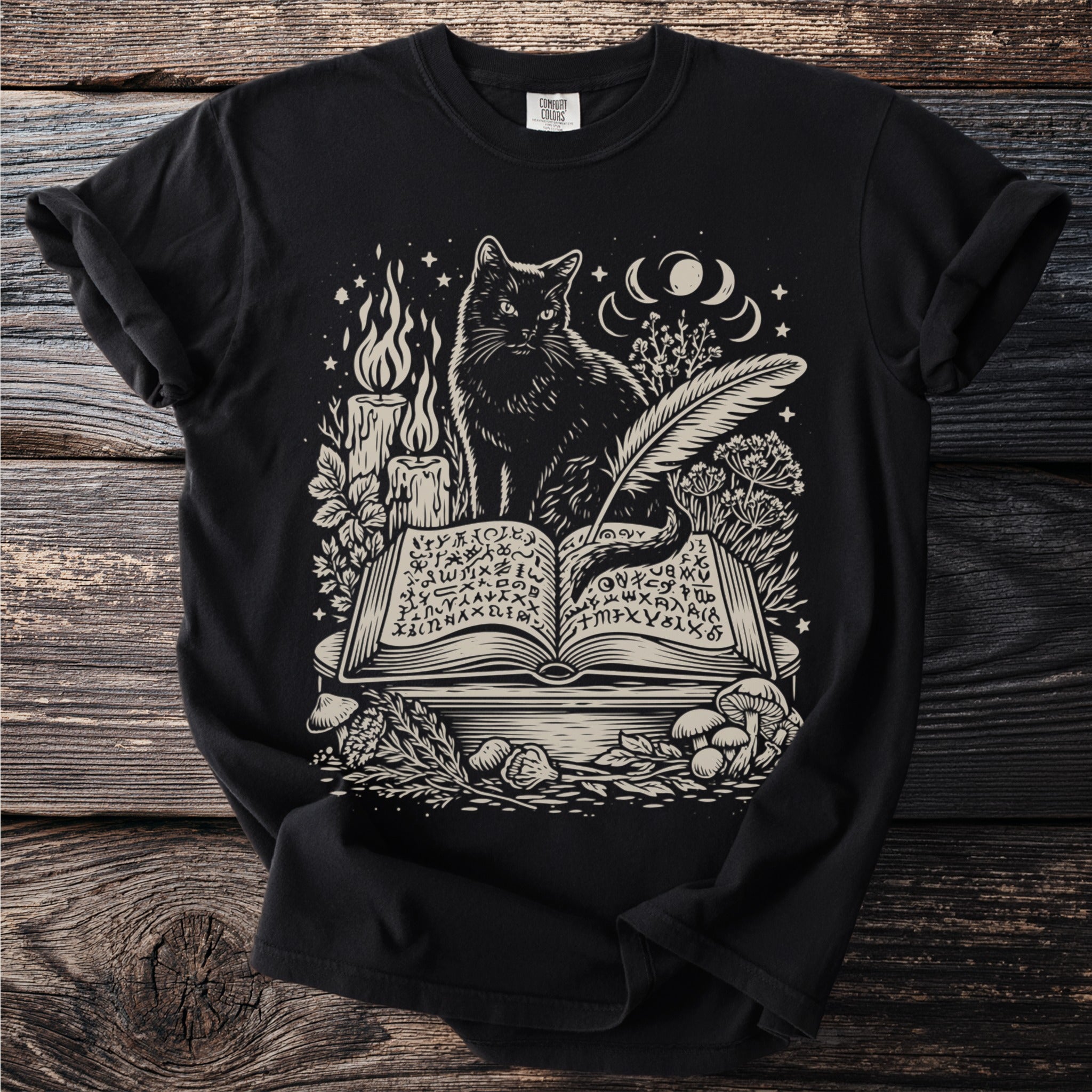 Black t-shirt with a cream graphic of a cat reading an open book with mystical symbols.