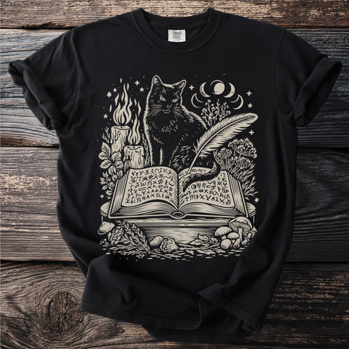 Black t-shirt with a cream graphic of a cat reading an open book with mystical symbols.