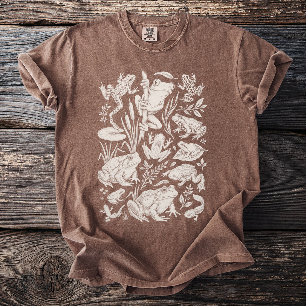 Brown t-shirt with an all-over illustration of various frogs, tadpoles, and aquatic plants.