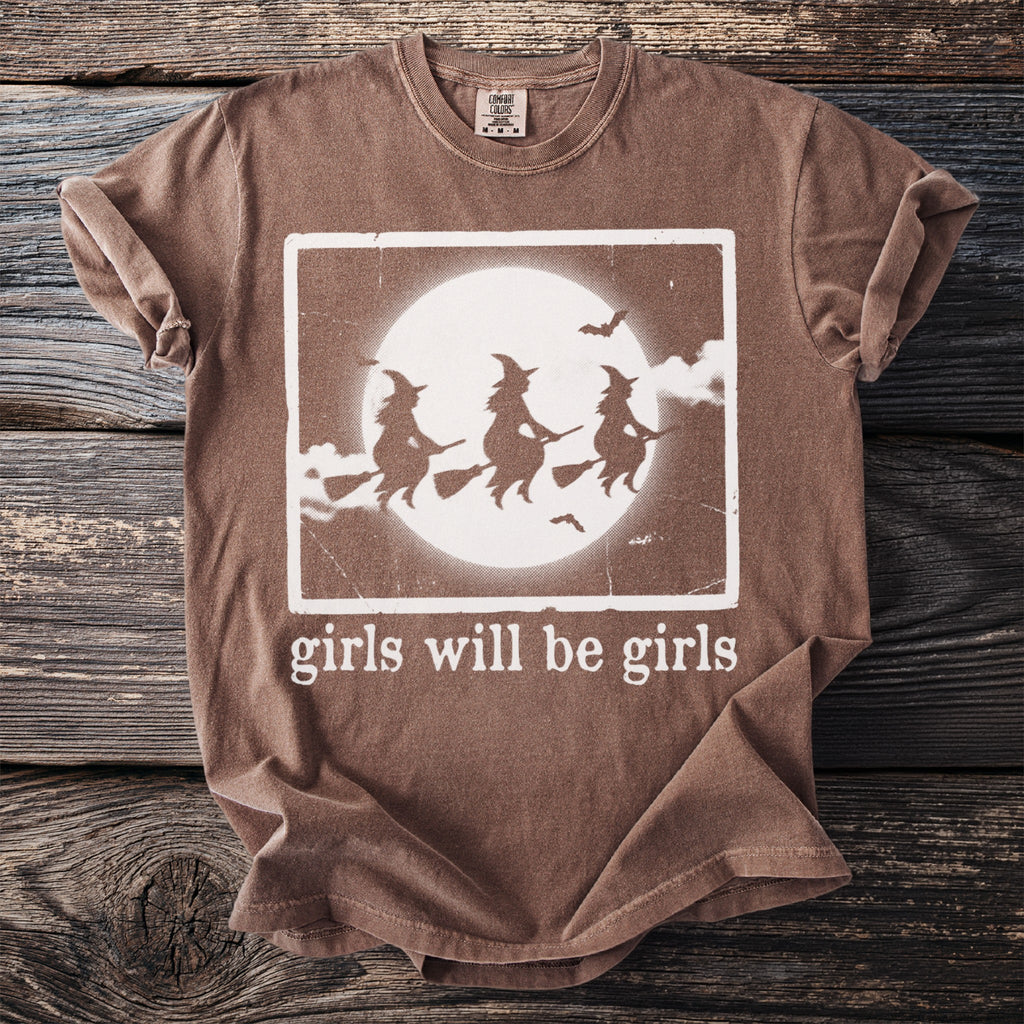 Brown t-shirt with white graphic of three witches flying and text "girls will be girls".