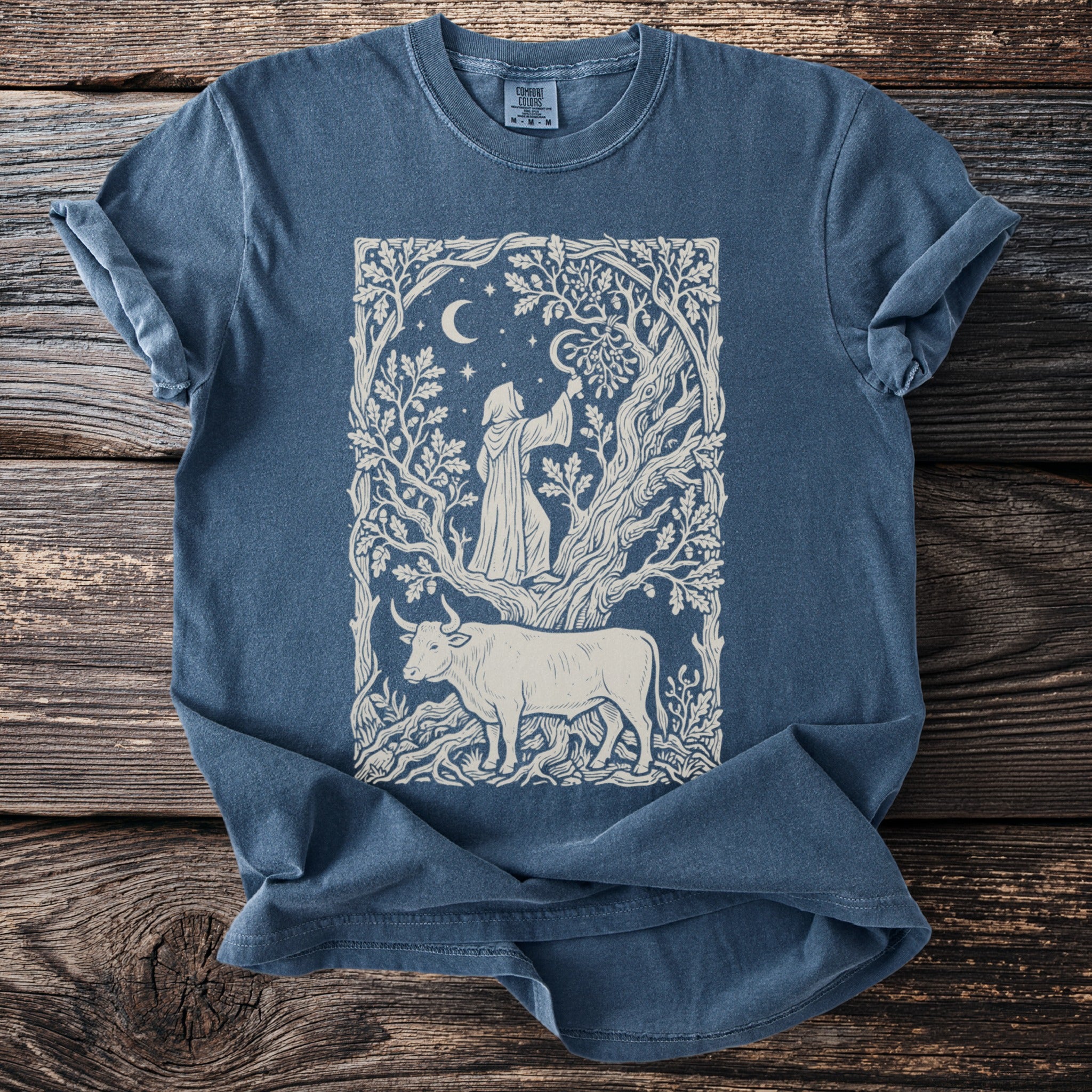 Blue t-shirt with a cream line art graphic of a hooded figure and a bull in a forest scene.