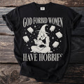 Black t-shirt with text "GOD FORBID WOMEN HAVE HOBBIFS" and witch reading amid books.