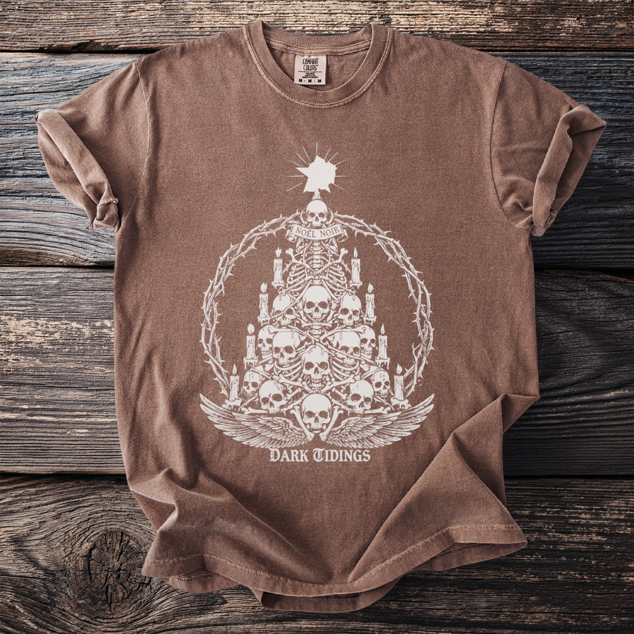 Brown t-shirt with a white graphic of a skull-and-bones Christmas tree and "DARK TIDINGS" text.