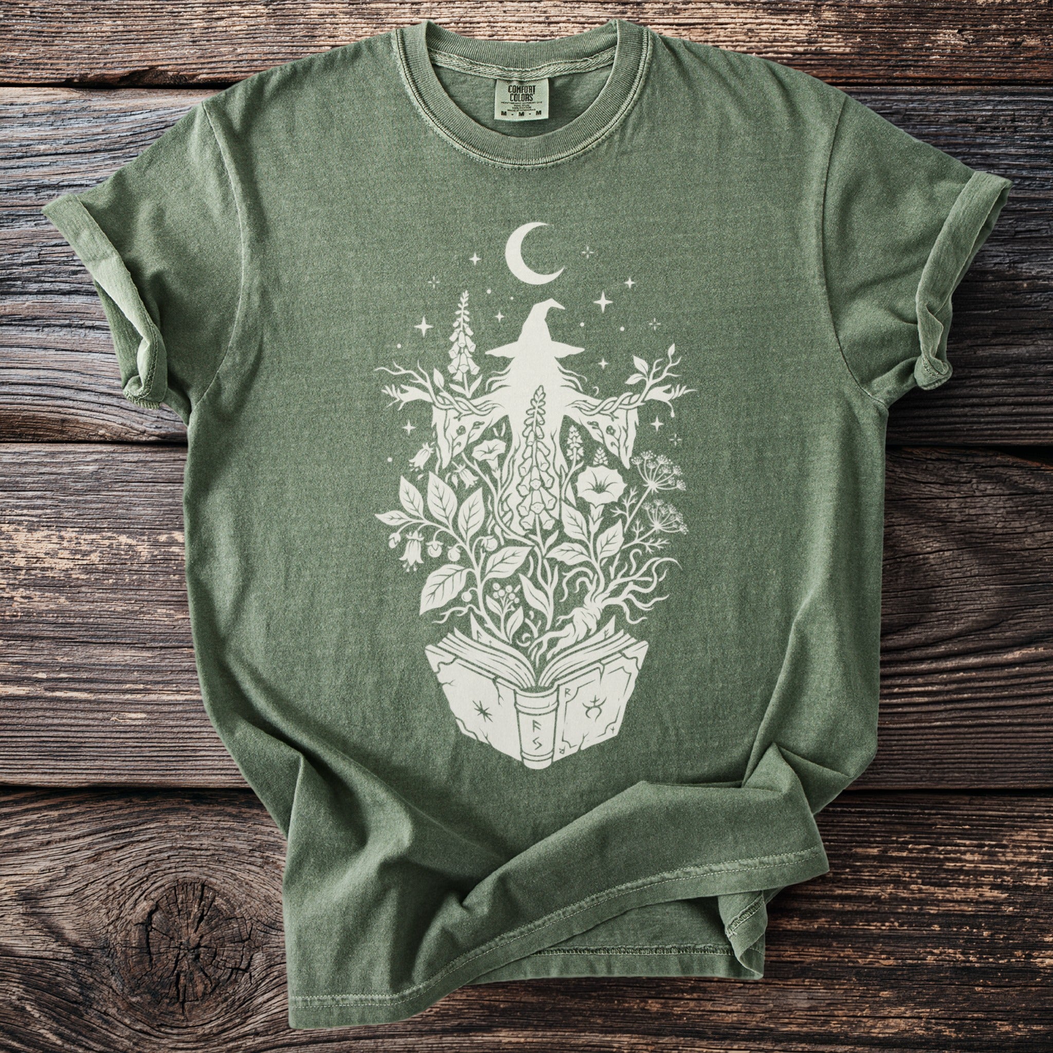 Green t-shirt with white witch and floral design emerging from an open book.