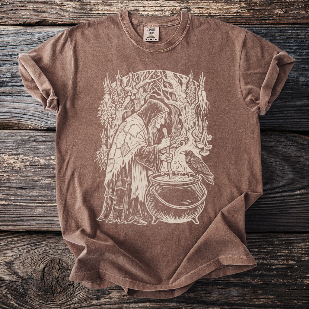 Brown t-shirt with a cream graphic of a witch stirring a cauldron with a raven.