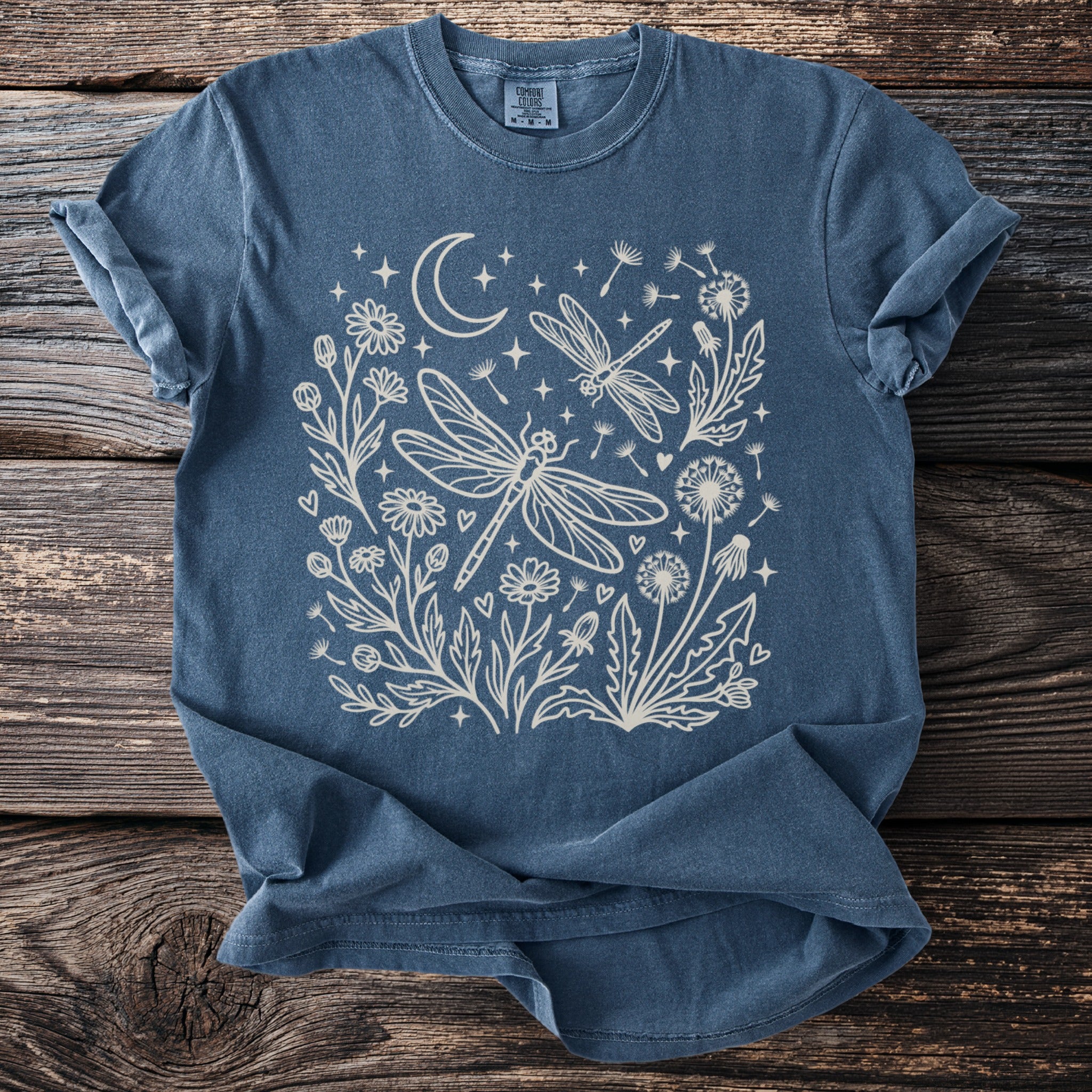 Blue t-shirt with a white line drawing of dragonflies, dandelions, flowers, moon, and stars.