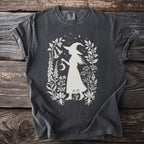 Dark t-shirt with white silhouette of a witch holding a crescent moon amongst botanical elements.