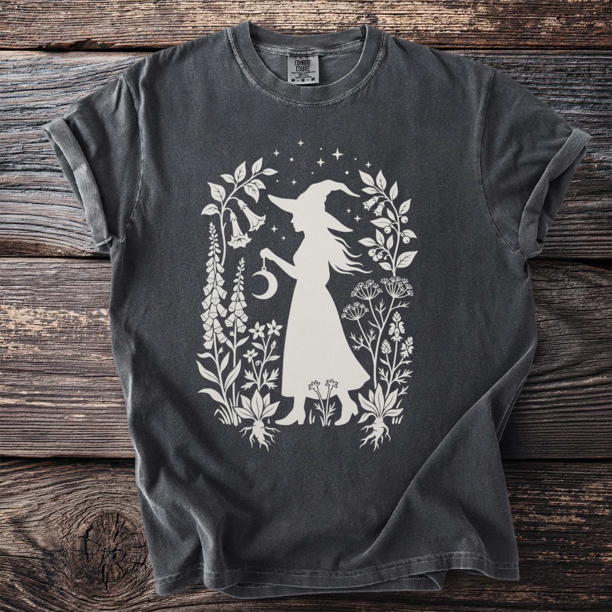 Dark t-shirt with white silhouette of a witch holding a crescent moon amongst botanical elements.