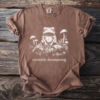 Brown t-shirt with a white graphic of a frog and mushrooms, text reads "currently decomposing".