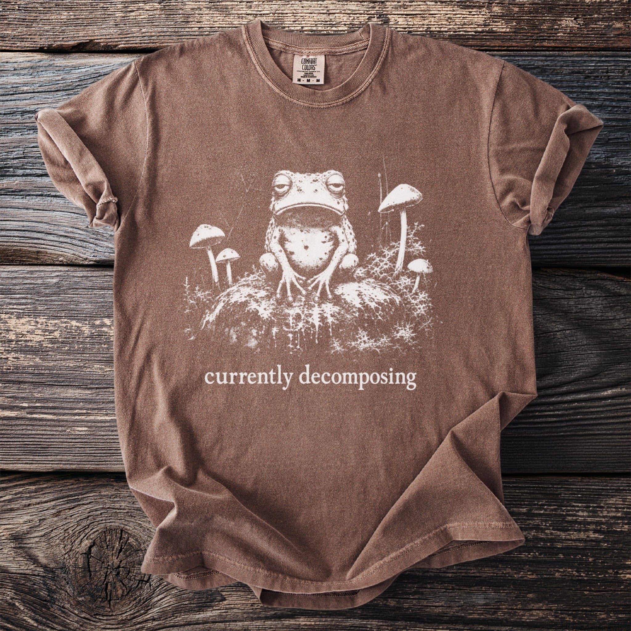 Brown t-shirt with a white graphic of a frog and mushrooms, text reads "currently decomposing".