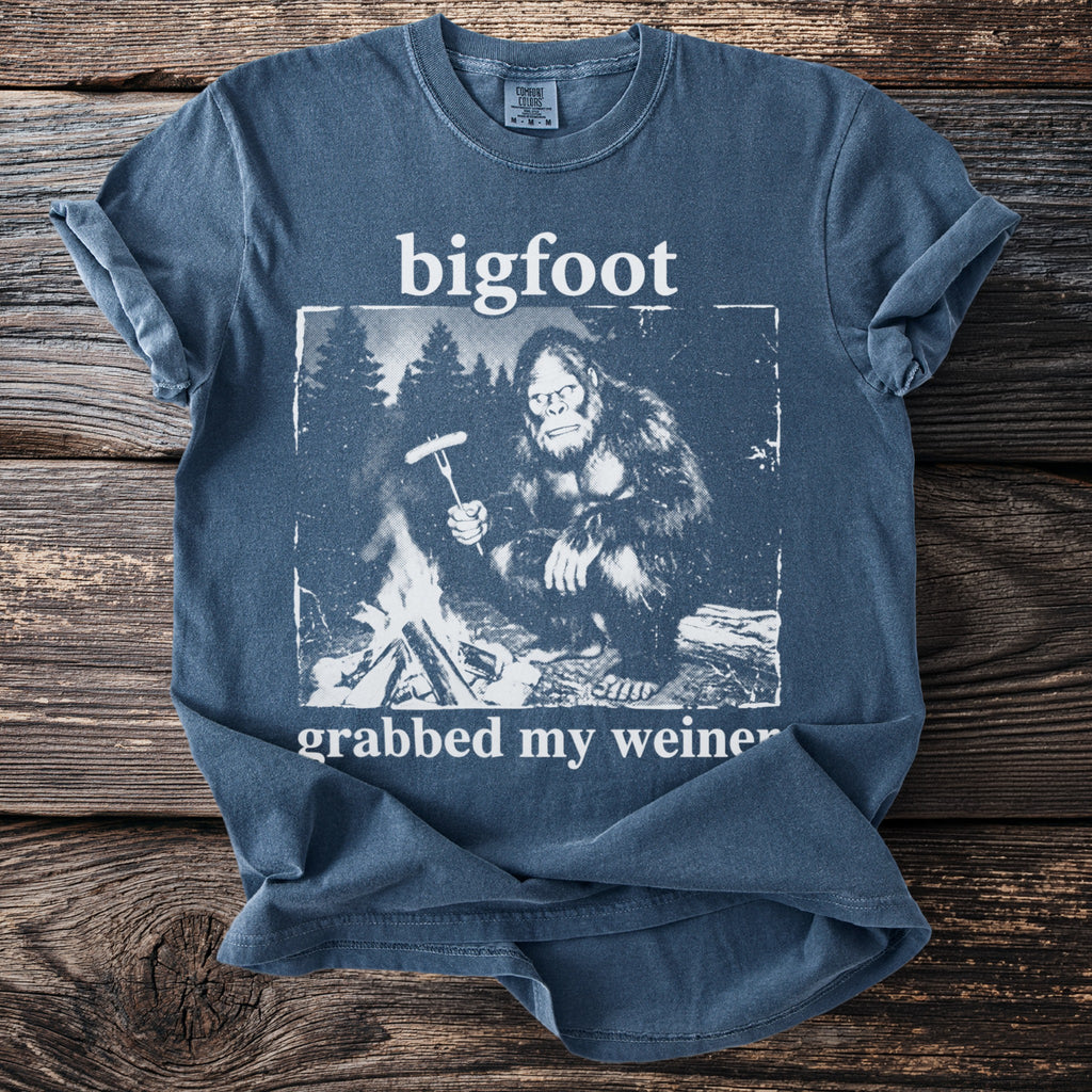 Blue t-shirt with graphic of Bigfoot roasting a hot dog over a campfire.