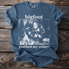 Blue t-shirt with graphic of Bigfoot roasting a hot dog over a campfire.