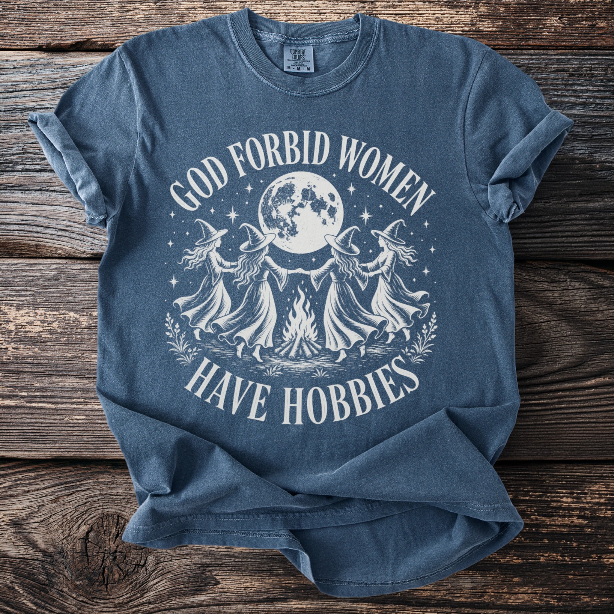 Blue t-shirt with white graphic of four witches dancing around a campfire under a full moon.