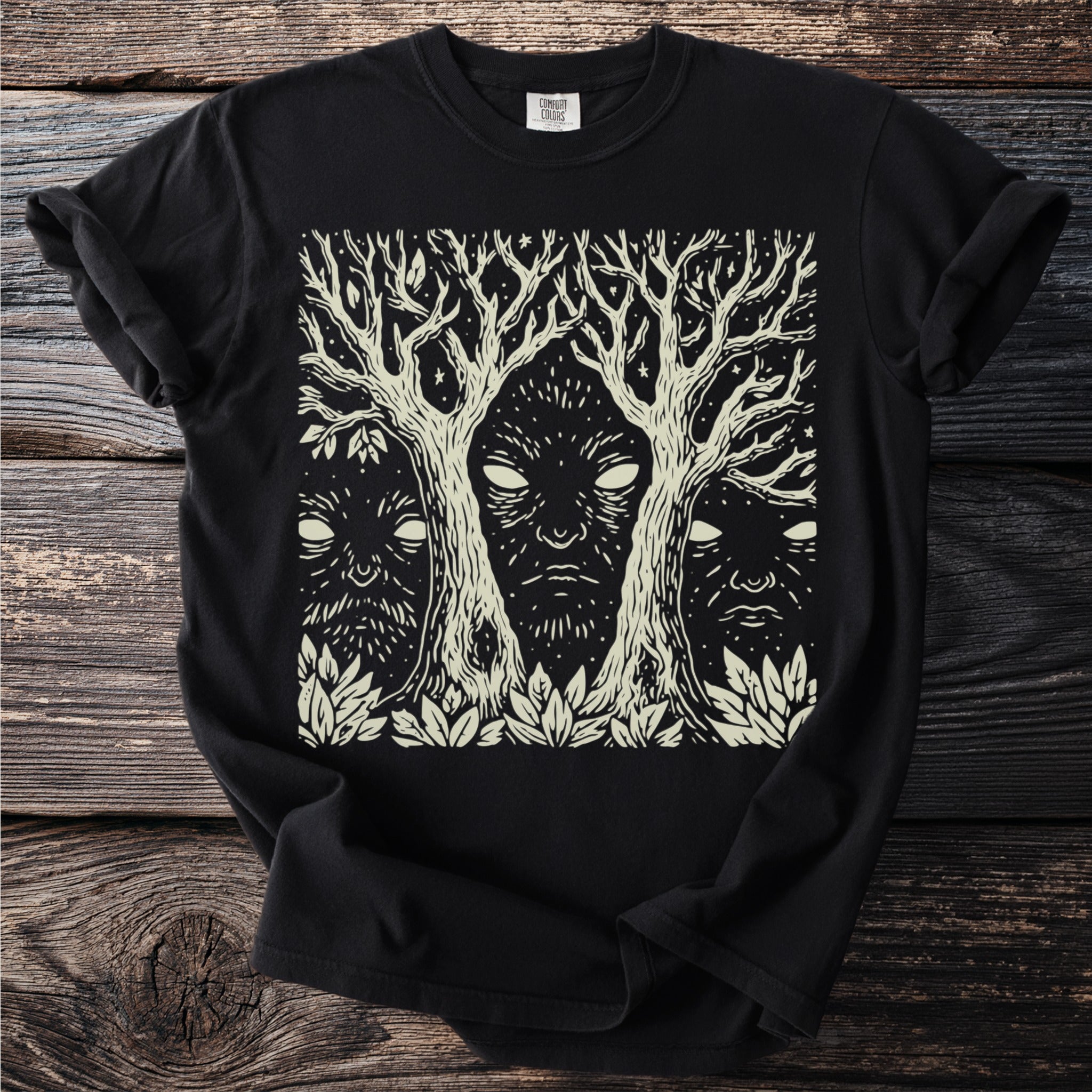 Black t-shirt with a cream linocut print of three faces within trees on a starry background.