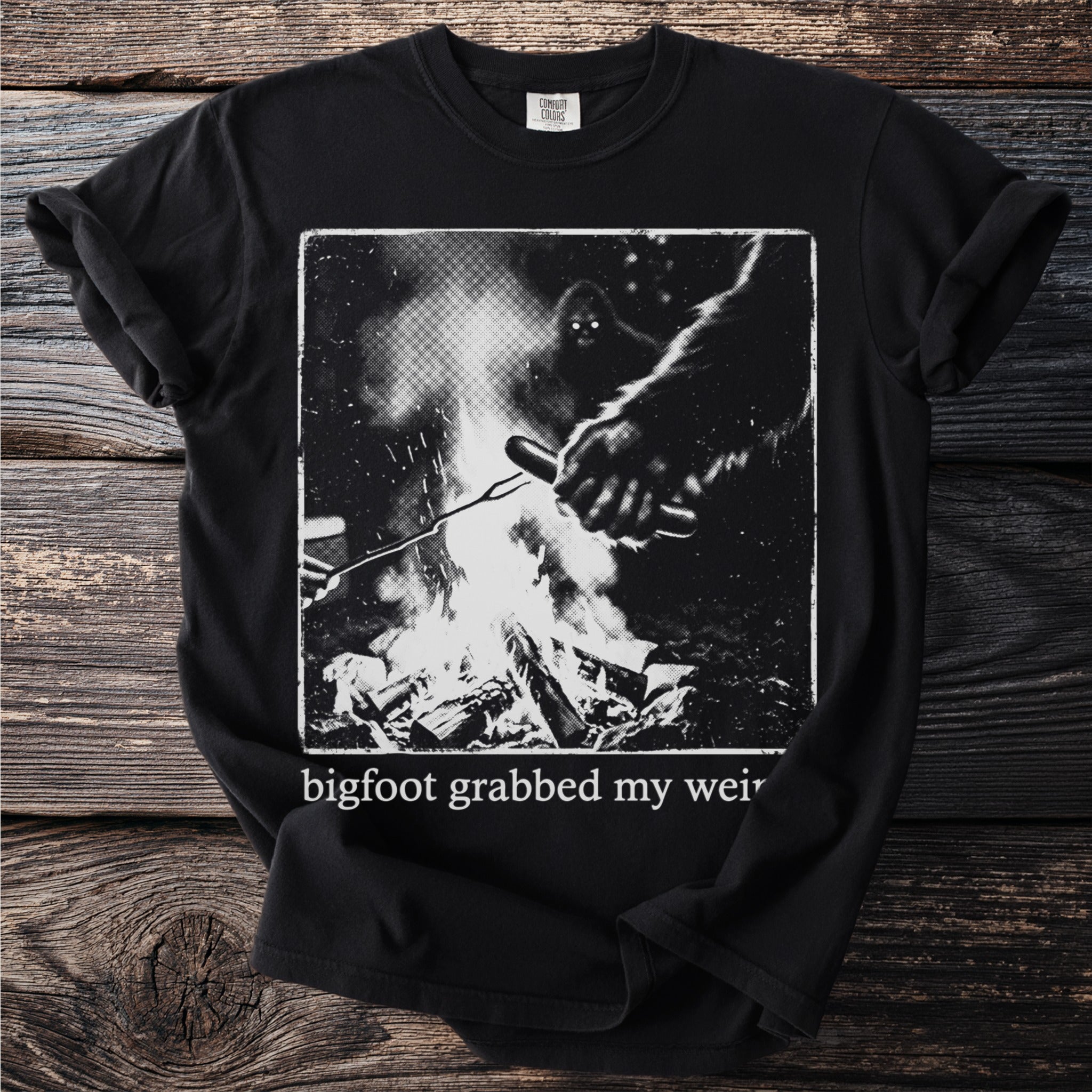 Black t-shirt with Bigfoot roasting a hot dog over a campfire graphic.