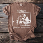 Brown t-shirt with white text and graphics of Bigfoot grabbing a stick.