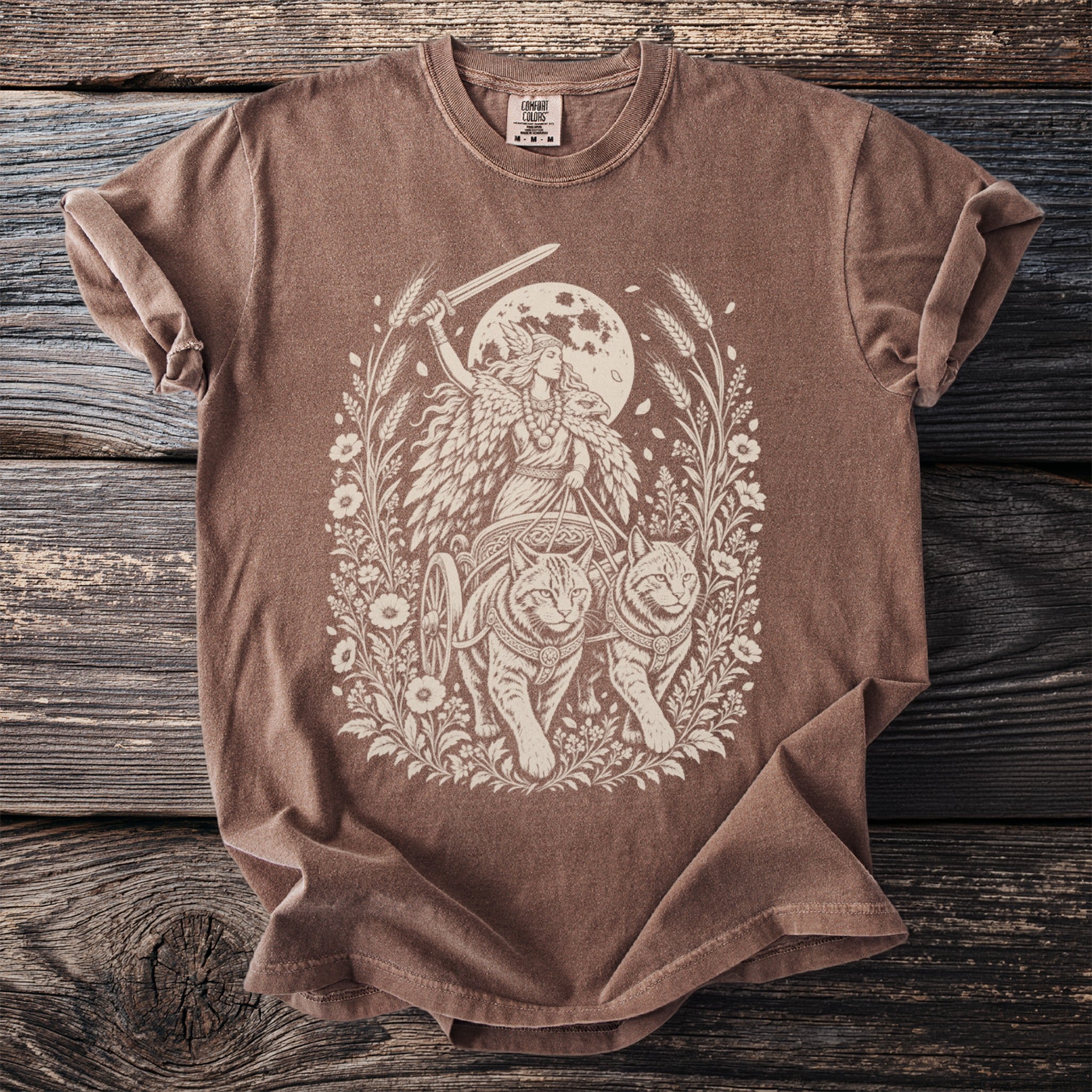 Brown t-shirt with a cream-colored illustration of a woman in a chariot pulled by two cats.