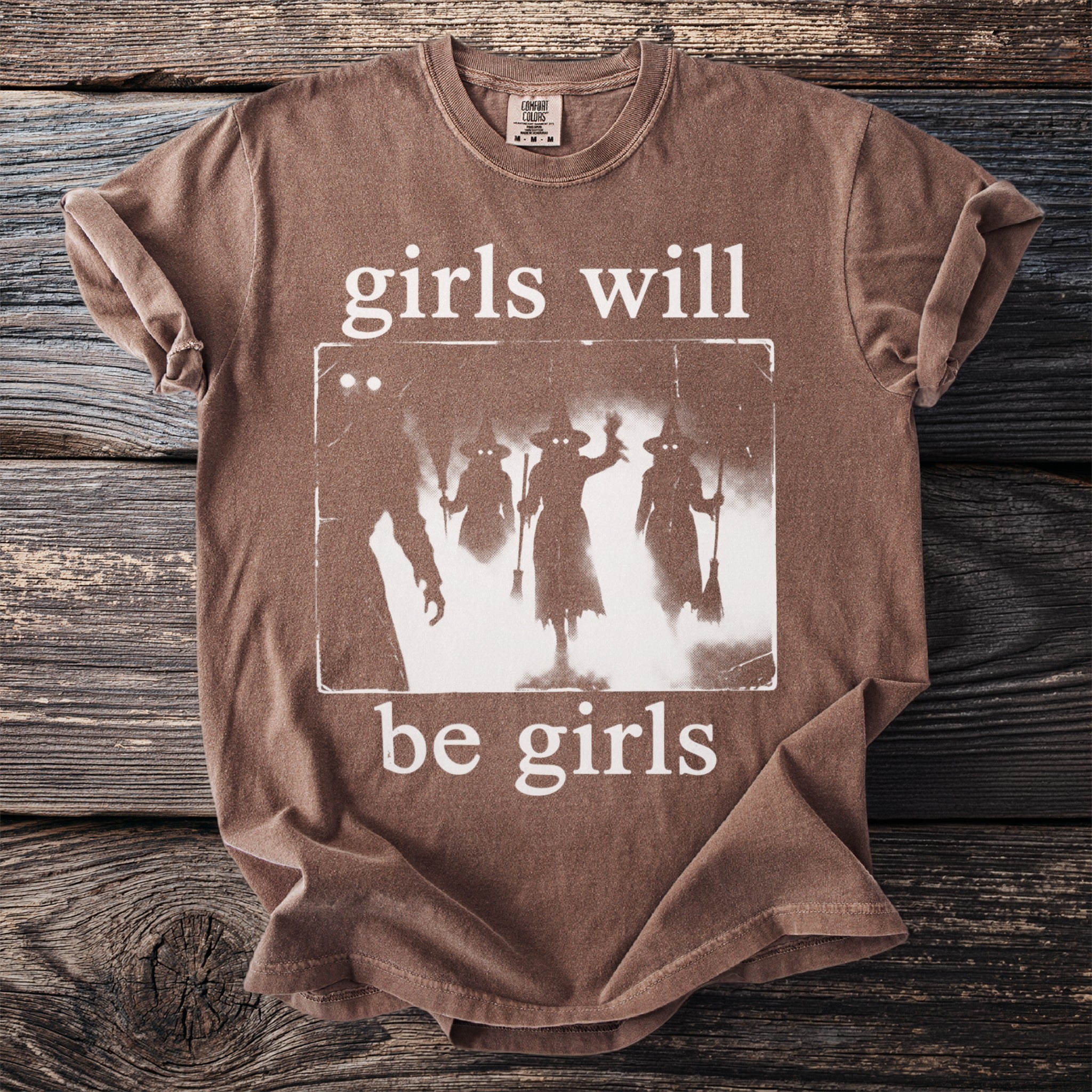 Brown t-shirt with glowing-eyed witches graphic and text "girls will be girls".