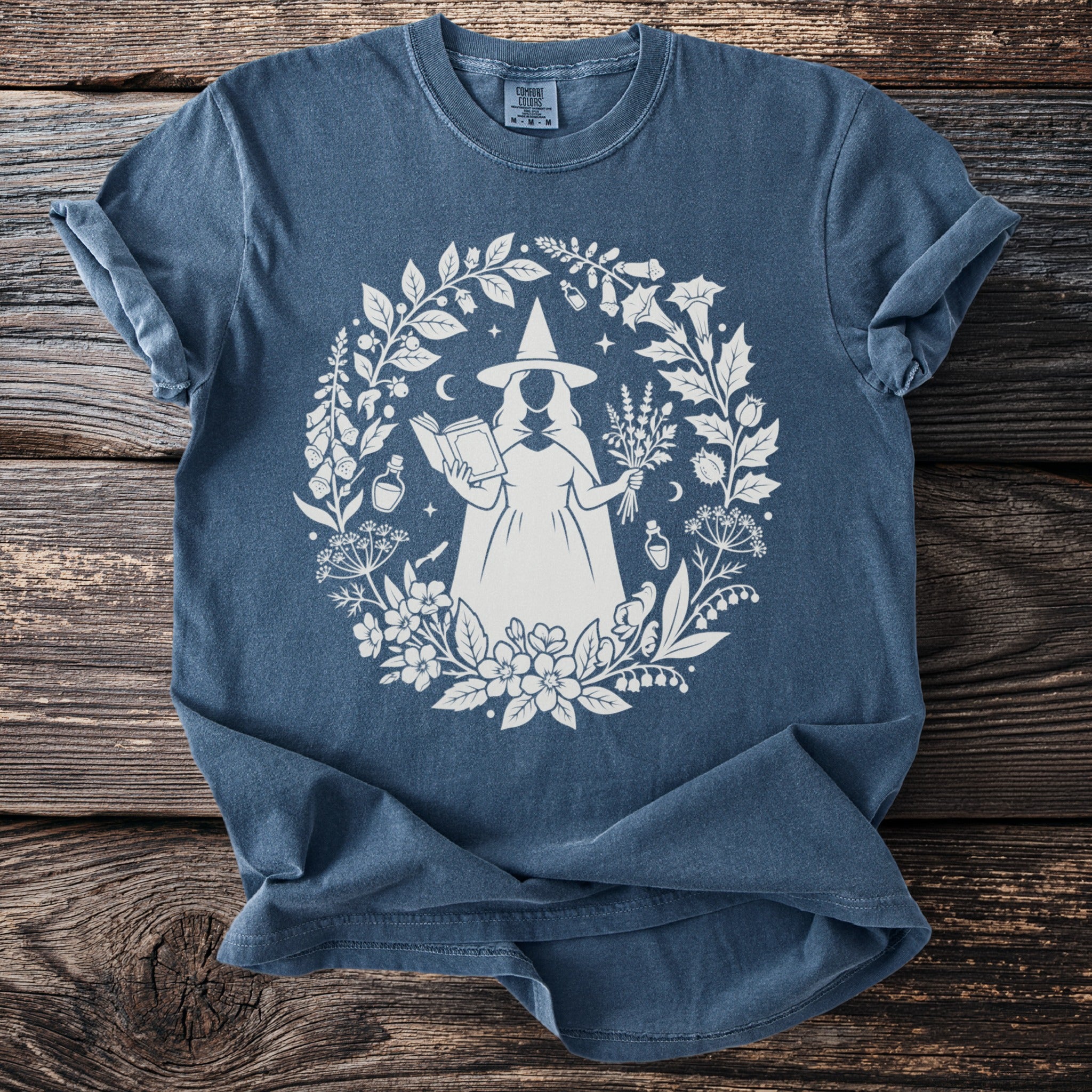 Blue t-shirt with circle design of a witch, book, herbs, and potions.