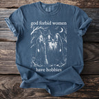 Blue t-shirt with witches, broom, lantern, moon, trees, and text "god forbid women have hobbies".